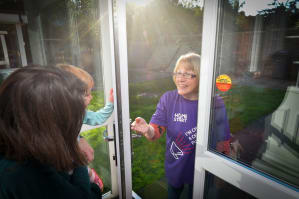Mum holding baby opens the door to her Home-Start volunteer, who is wearing a Home-Start T-Shirt